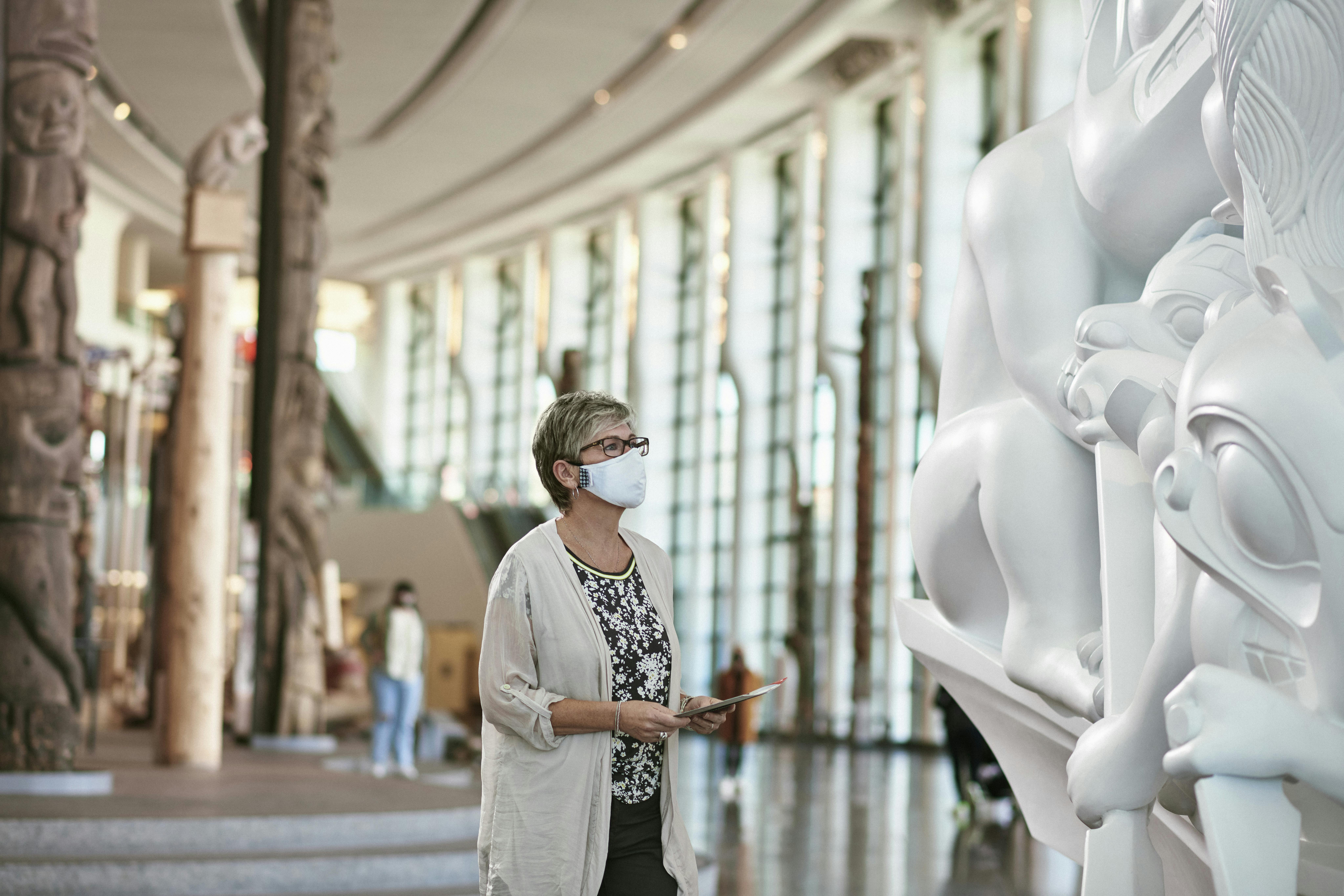 一位戴着面具的女士在一个宽敞的现代画廊里观察到一个大型雕塑，画廊的天花板很高，窗户很大。