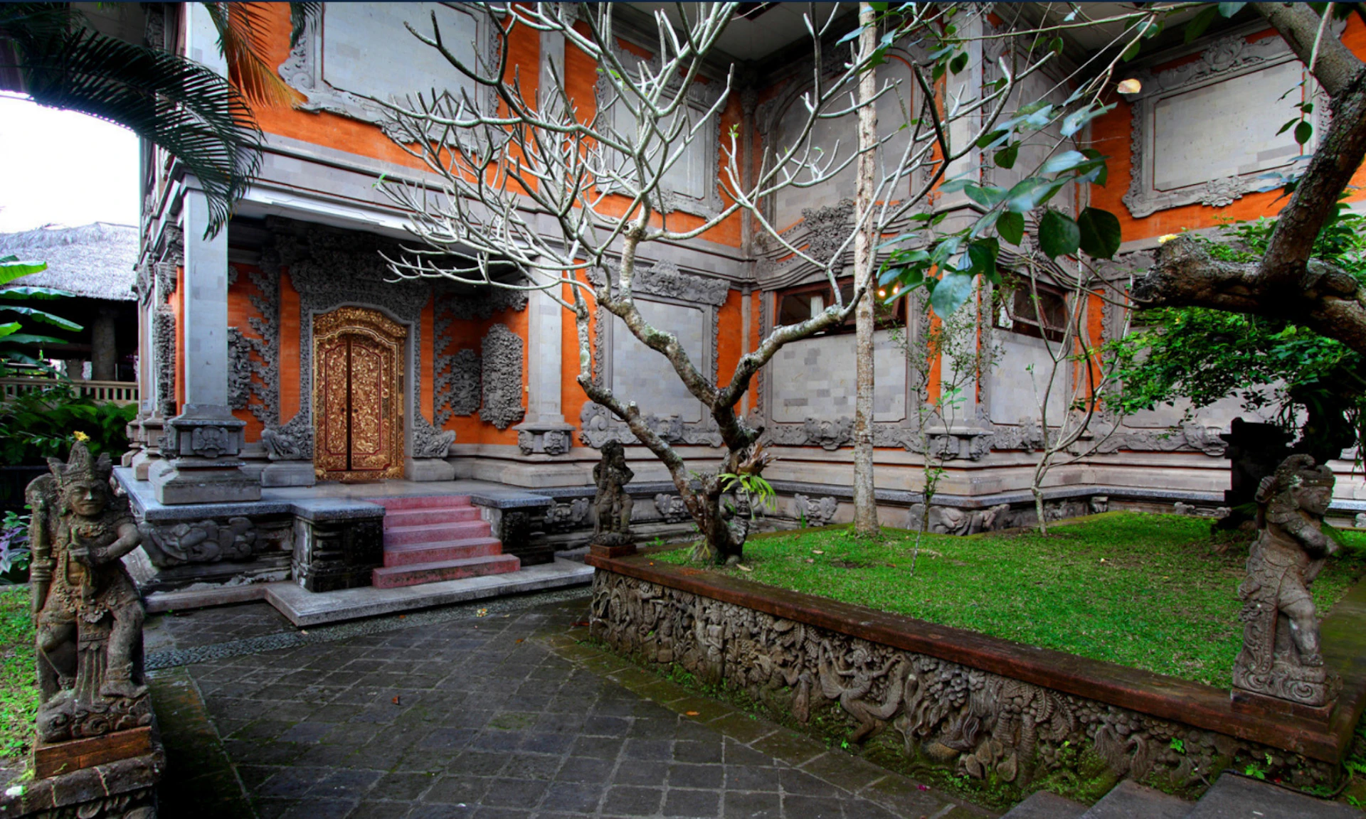 A traditional courtyard with a decorative wooden door, stone carvings, a tree, grass, and paved flooring.