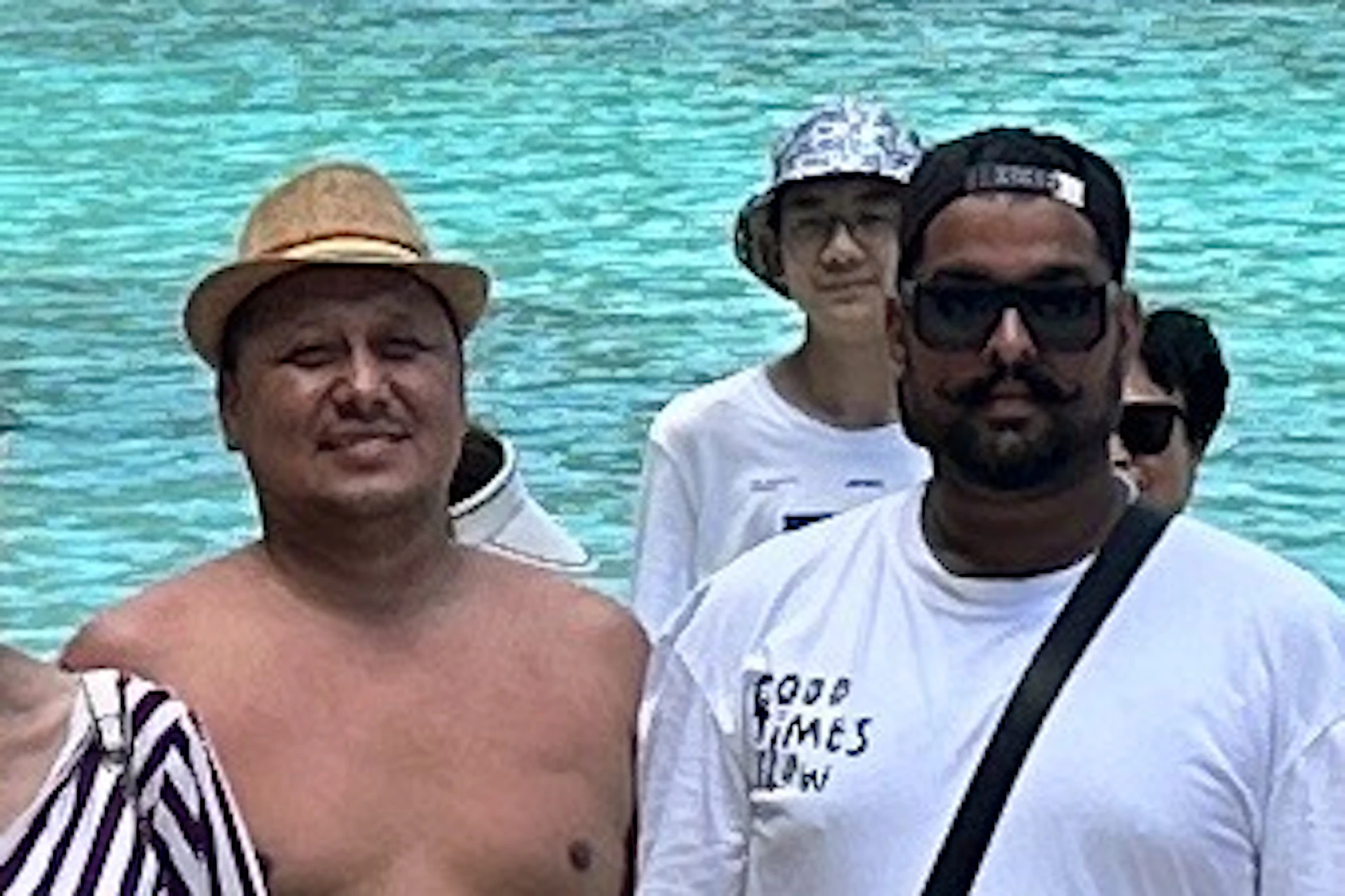 A group of people stands on a beach with clear blue water and rocky green cliffs in the background.