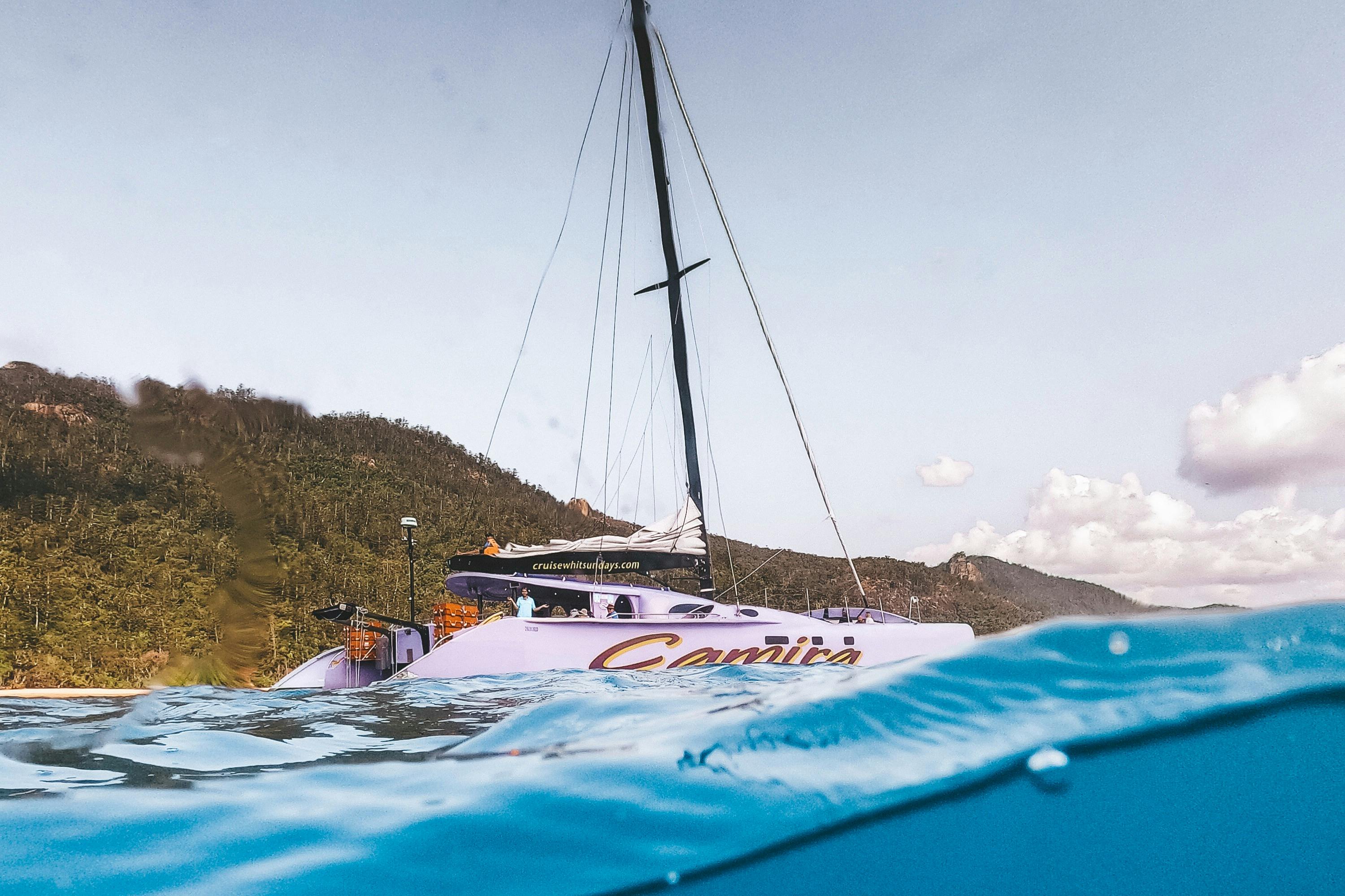 Парусник под названием «Камира» стоит на якоре в спокойной воде недалеко от лесистой береговой линии, а его мачта простирается до частично облачного неба.