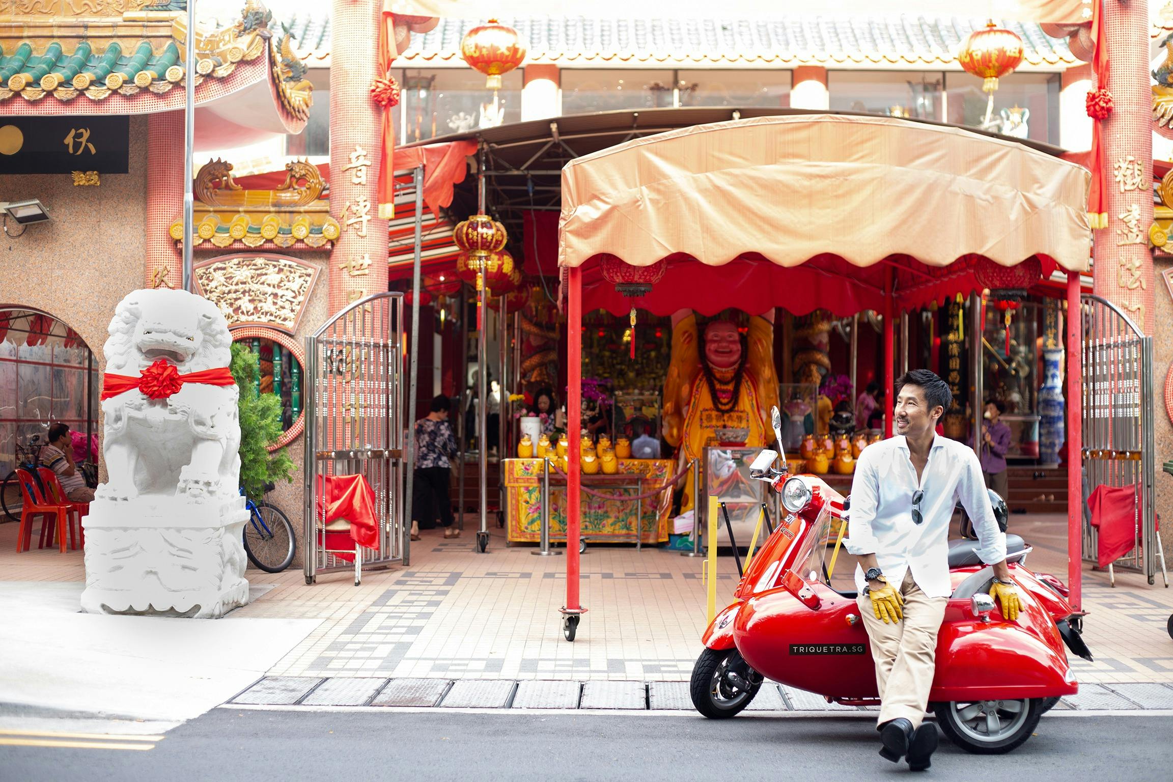 Un homme vêtu d'une chemise blanche et d'un pantalon beige s'appuie sur un scooter rouge devant un étal de marché coloré avec des auvents et des lanternes rouges.