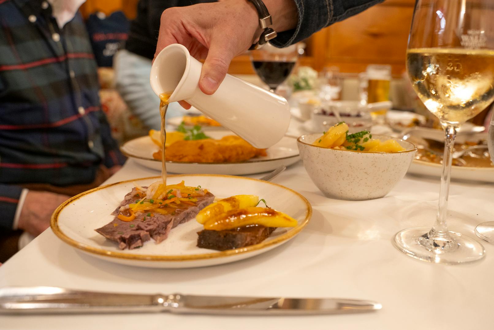 Uma mão deita molho de um jarro branco para um prato de comida numa mesa de jantar, com pratos e bebidas ao fundo.