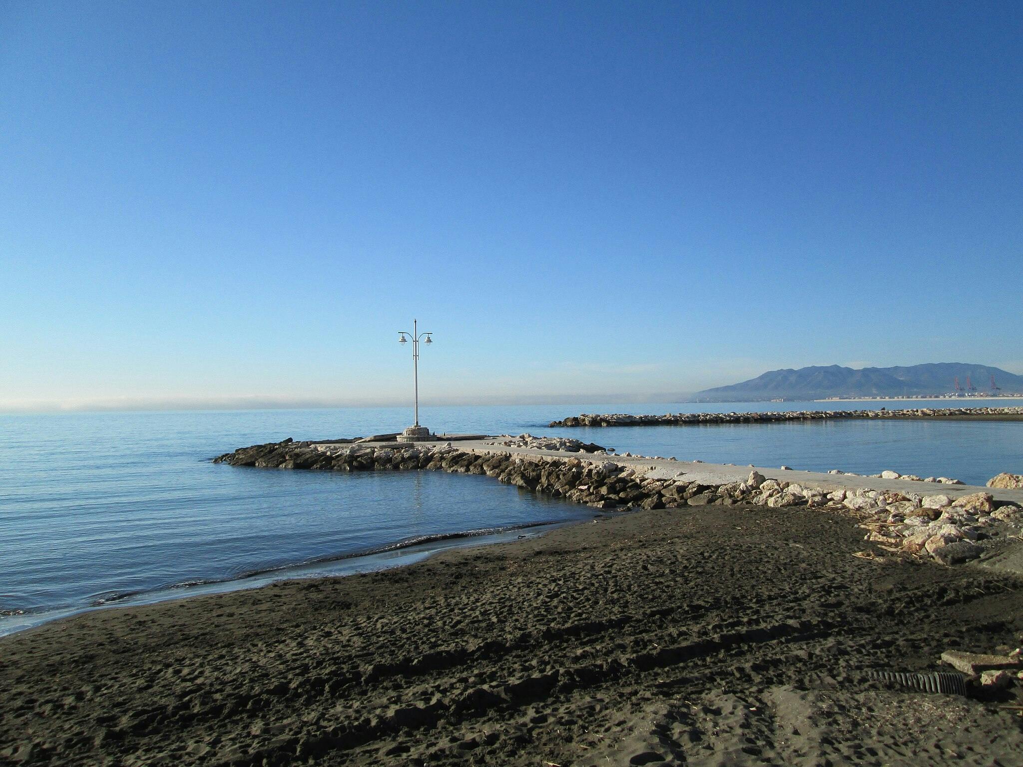Rocky pier extending into calm sea with two street lamps, sandy beach in foreground, and distant mountains under clear blue sky.