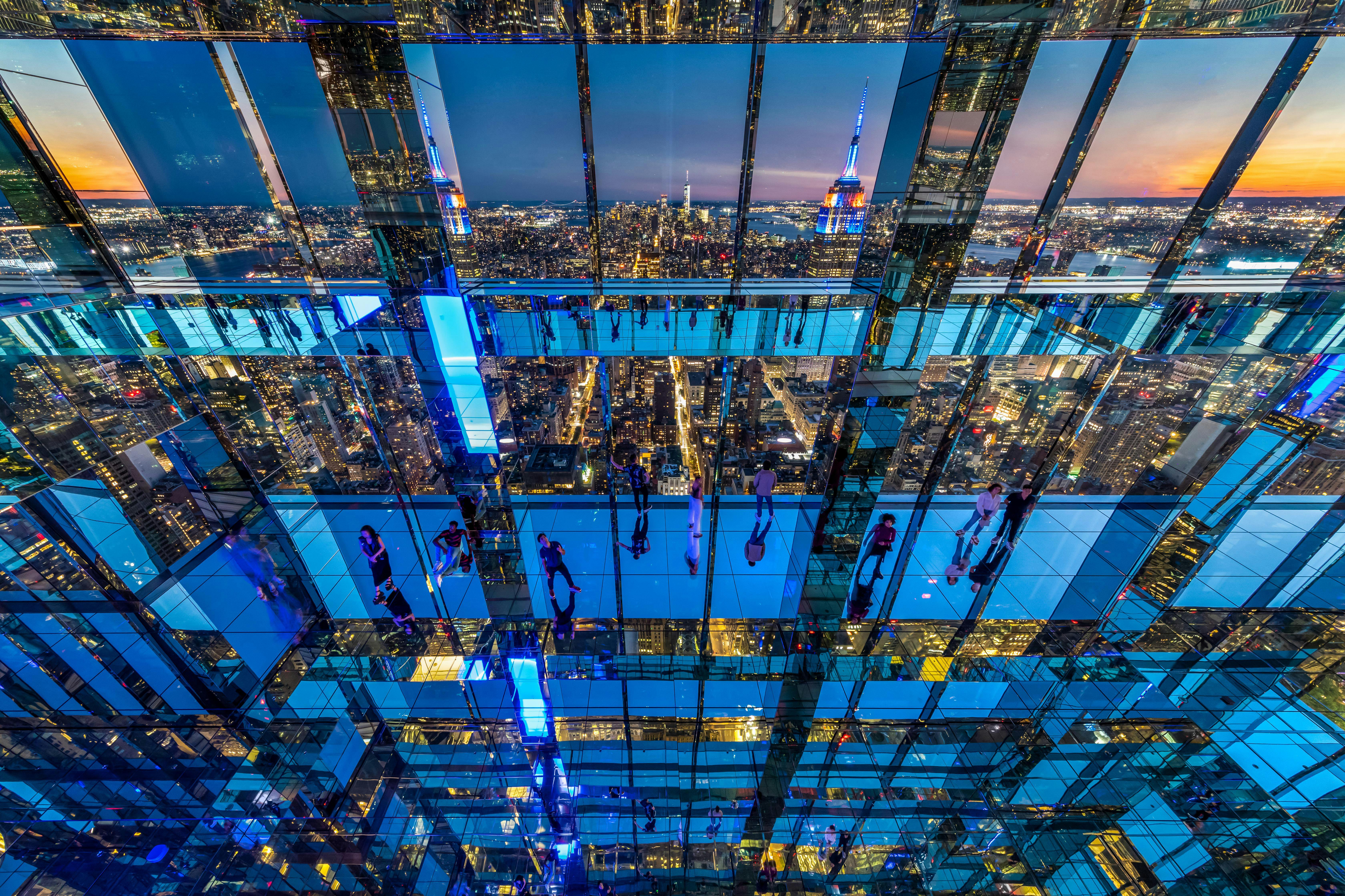 People observing a cityscape from within a glass-walled structure with multiple reflections and neon blue lighting.
