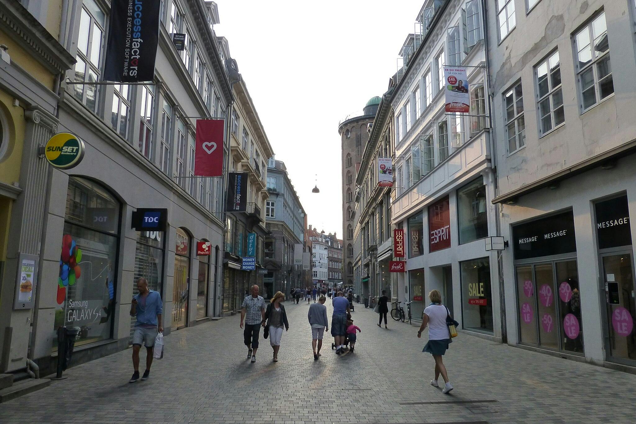 Οι άνθρωποι περπατούν κατά μήκος ενός πλακόστρωτου δρόμου που πλαισιώνεται από κτίρια με πινακίδες καταστημάτων, πανό και μεγάλα παράθυρα.