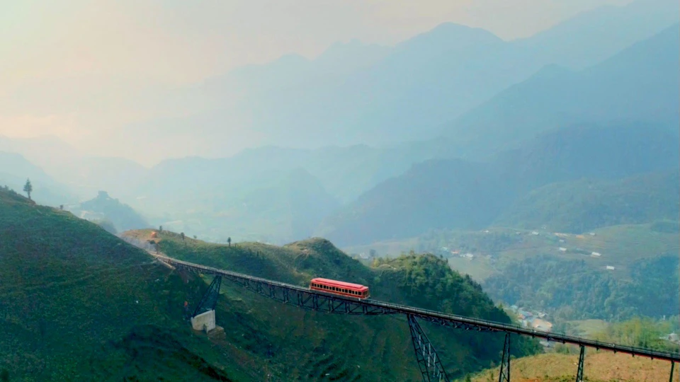 A red train crosses a tall bridge over a green hilly landscape with misty mountains in the background.