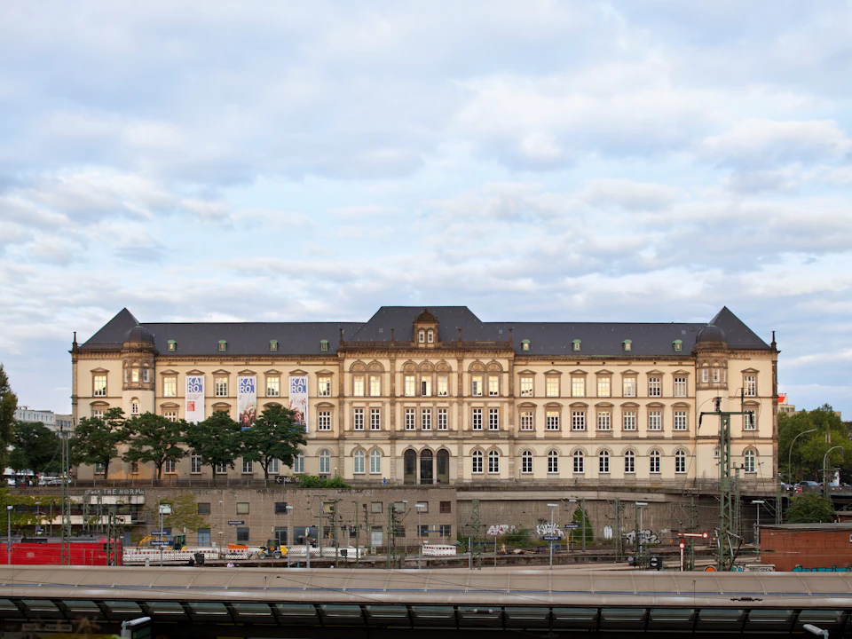 A large, historic building with numerous windows and decorative architectural details stands under a partly cloudy sky.