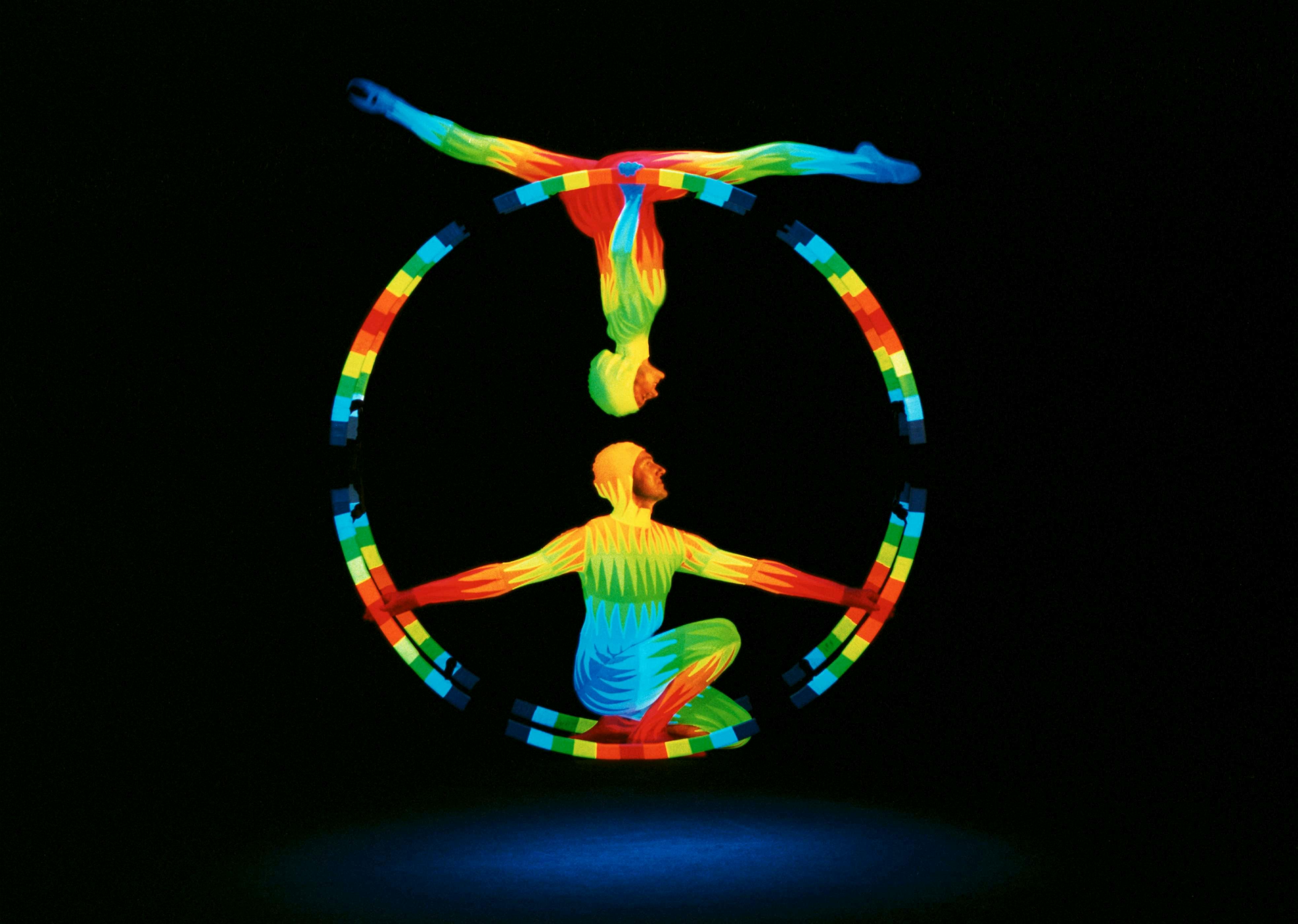 Two performers in colorful, glowing outfits balance on a circular frame in a dark setting, one upside down above the other.