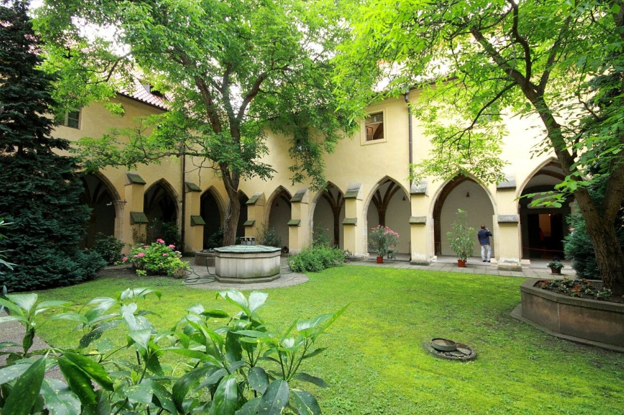Ein friedlicher Innenhof mit Grünflächen, Bäumen, einem zentralen Brunnen und einem bogenförmigen Weg entlang eines gelben Gebäudes. Eine Person ist zu sehen.