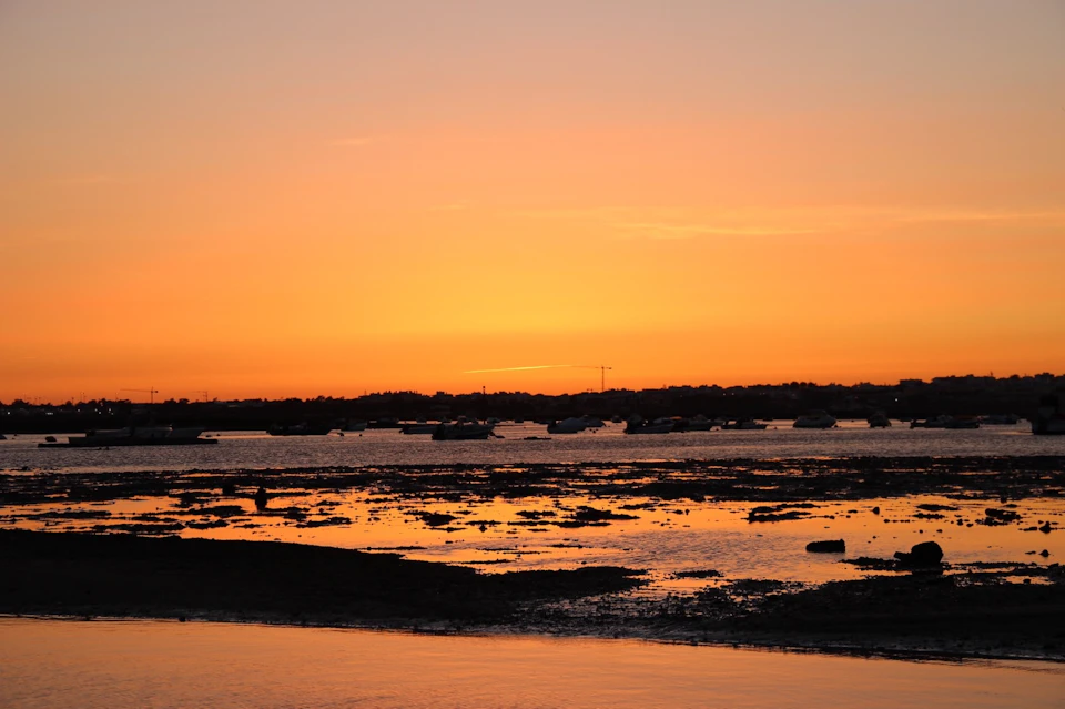 A vibrant sunset over a coastal area with boats on the water and reflections on the wet sand during low tide