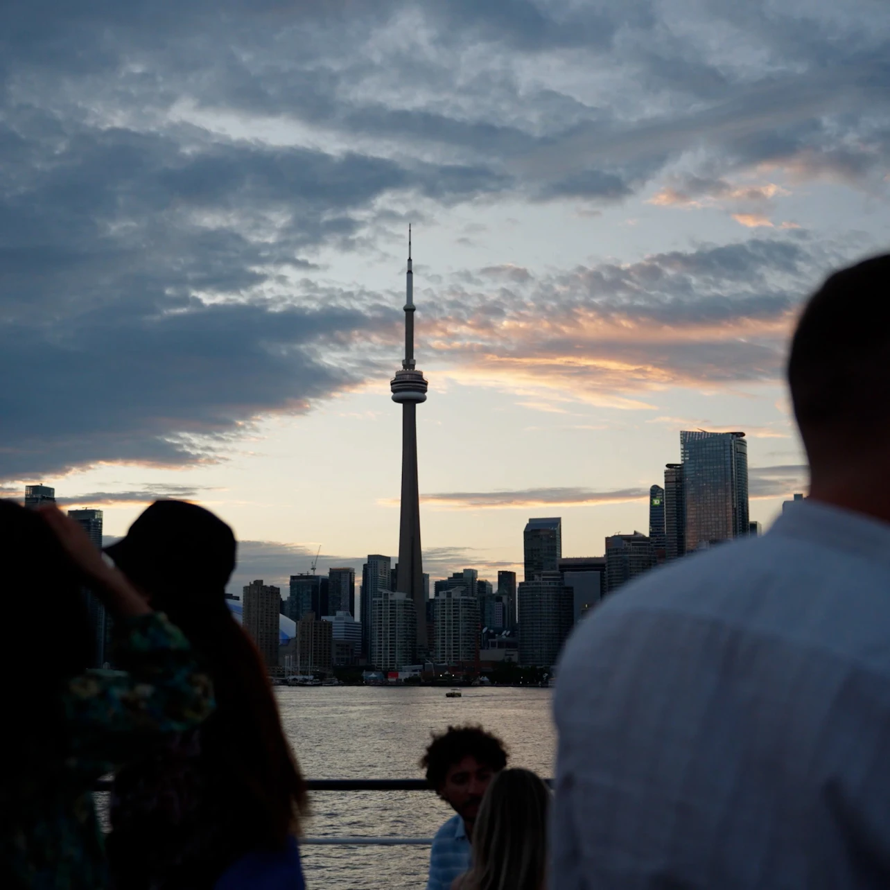 A city skyline with the CN Tower at sunset, seen from a waterfront area with silhouettes of people in the foreground.