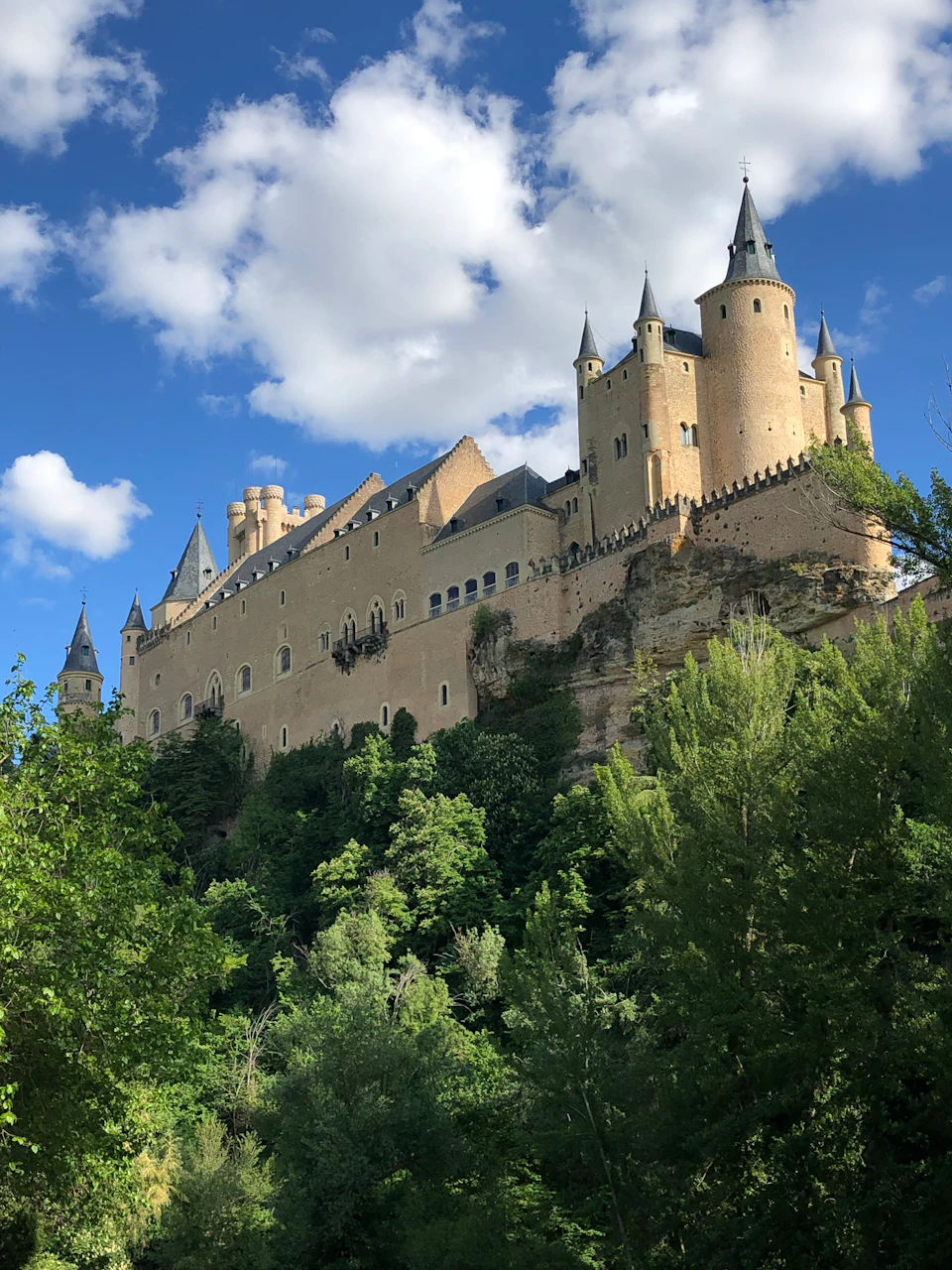 A large medieval castle with multiple turrets perched on a hilltop, surrounded by lush greenery and under a cloudy blue sky.