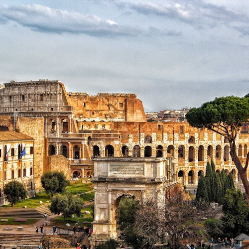 Il Colosseo a Roma con l'architettura storica circostante, gli alberi e il cielo nuvoloso sullo sfondo.