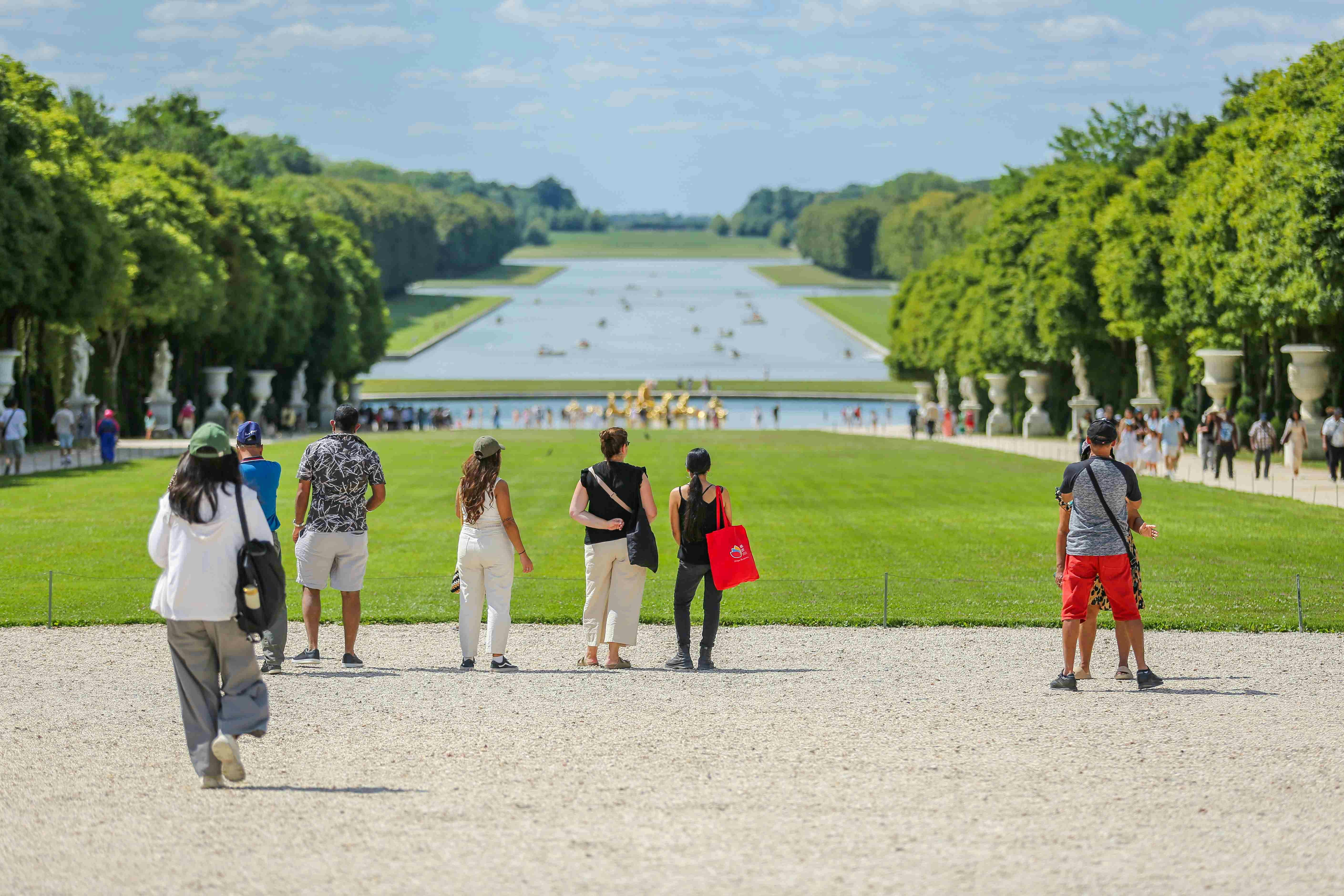 People walking towards a large garden with manicured lawns, fountains, and a long reflecting pool, bordered by trees.