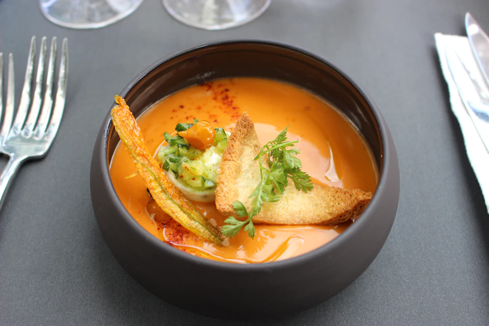 A bowl of orange soup garnished with a stuffed zucchini flower, herbs, and a piece of toasted bread.