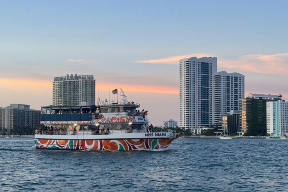 A colorful boat named "Miss Miami" on water with tall buildings and a sunset sky in the background.