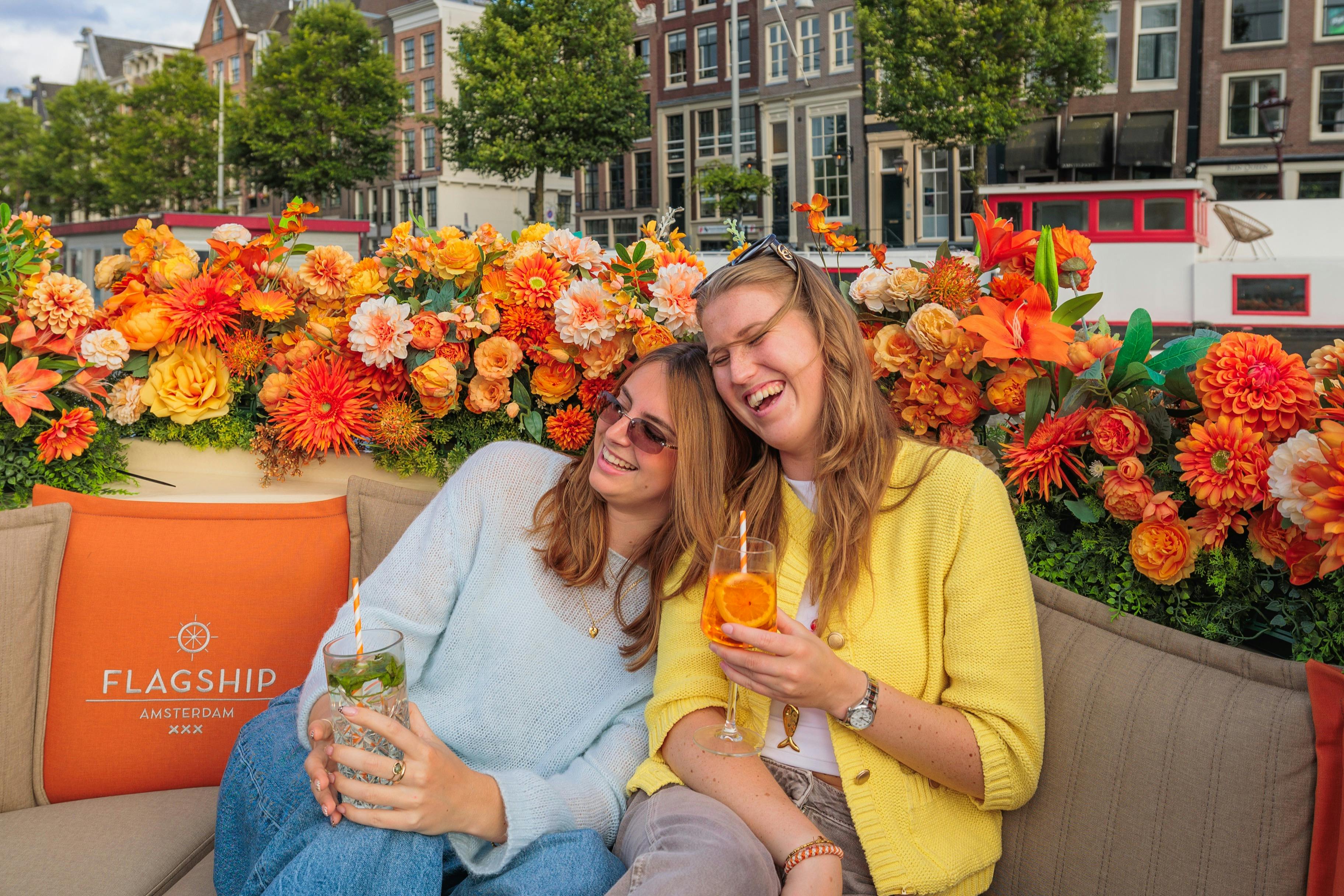 Zwei lächelnde Frauen mit Getränken in der Hand sitzen auf einem Sofa, im Hintergrund sind leuchtend orangefarbene Blumen und eine Stadtkulisse zu sehen.
