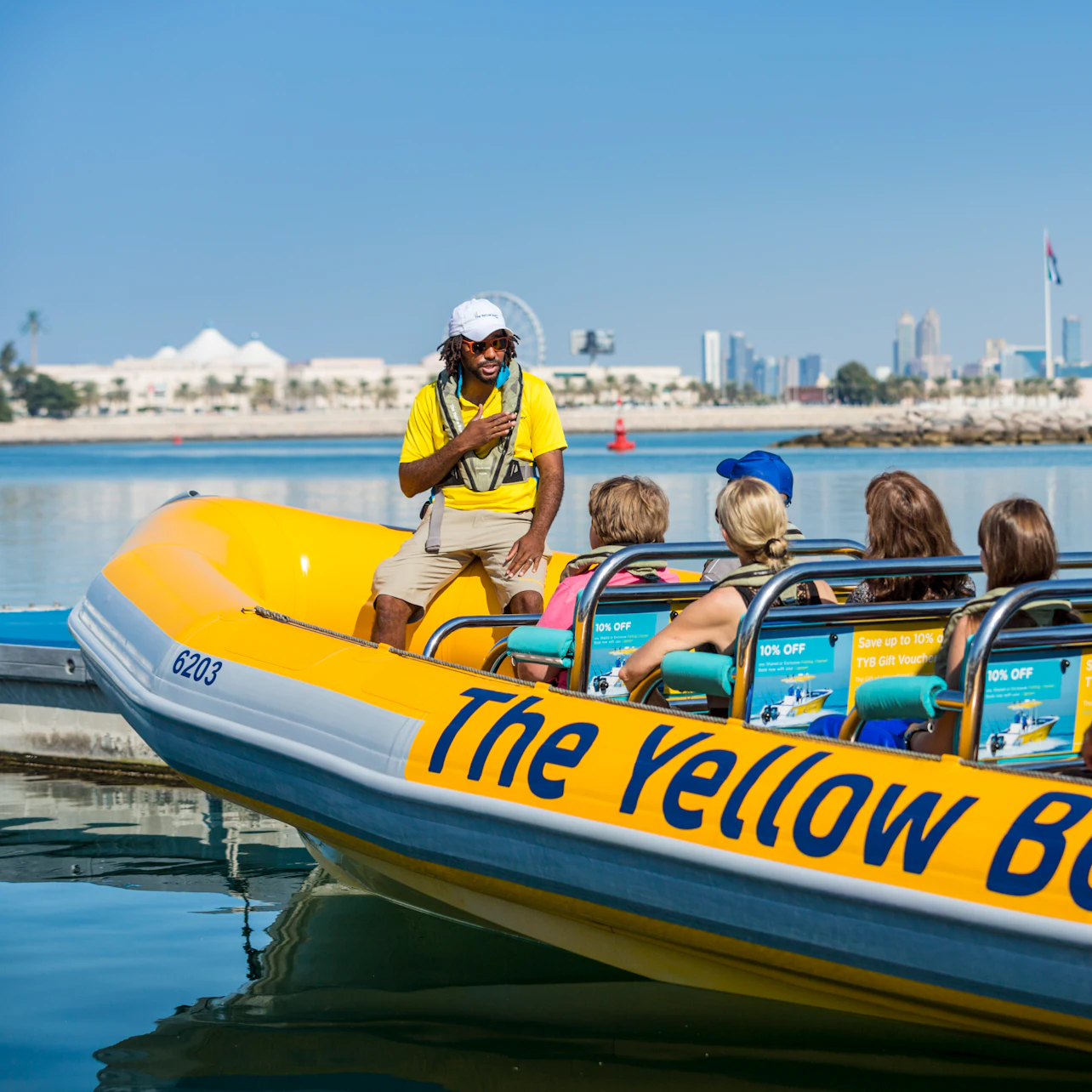 A tour guide in a yellow shirt and white hat stands on a yellow boat named "The Yellow Boat," talking to seated passengers.