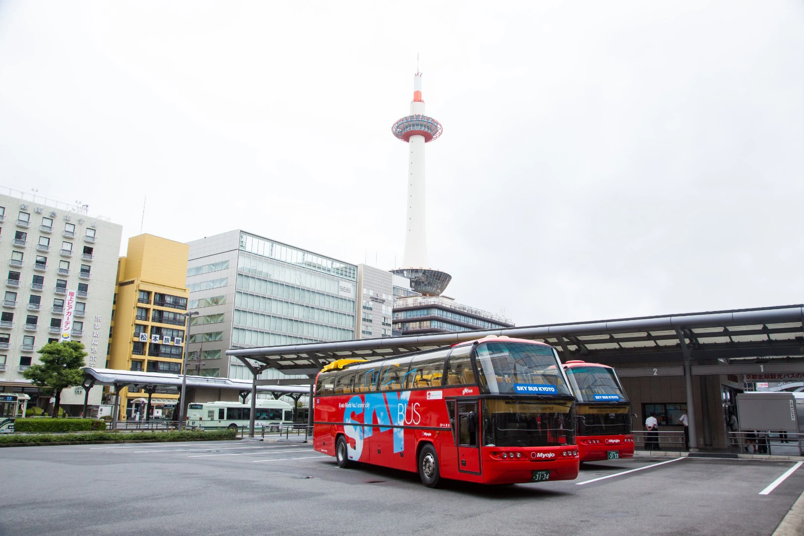A modern bus terminal with red tour buses, a tall tower in the background, and surrounding urban buildings.