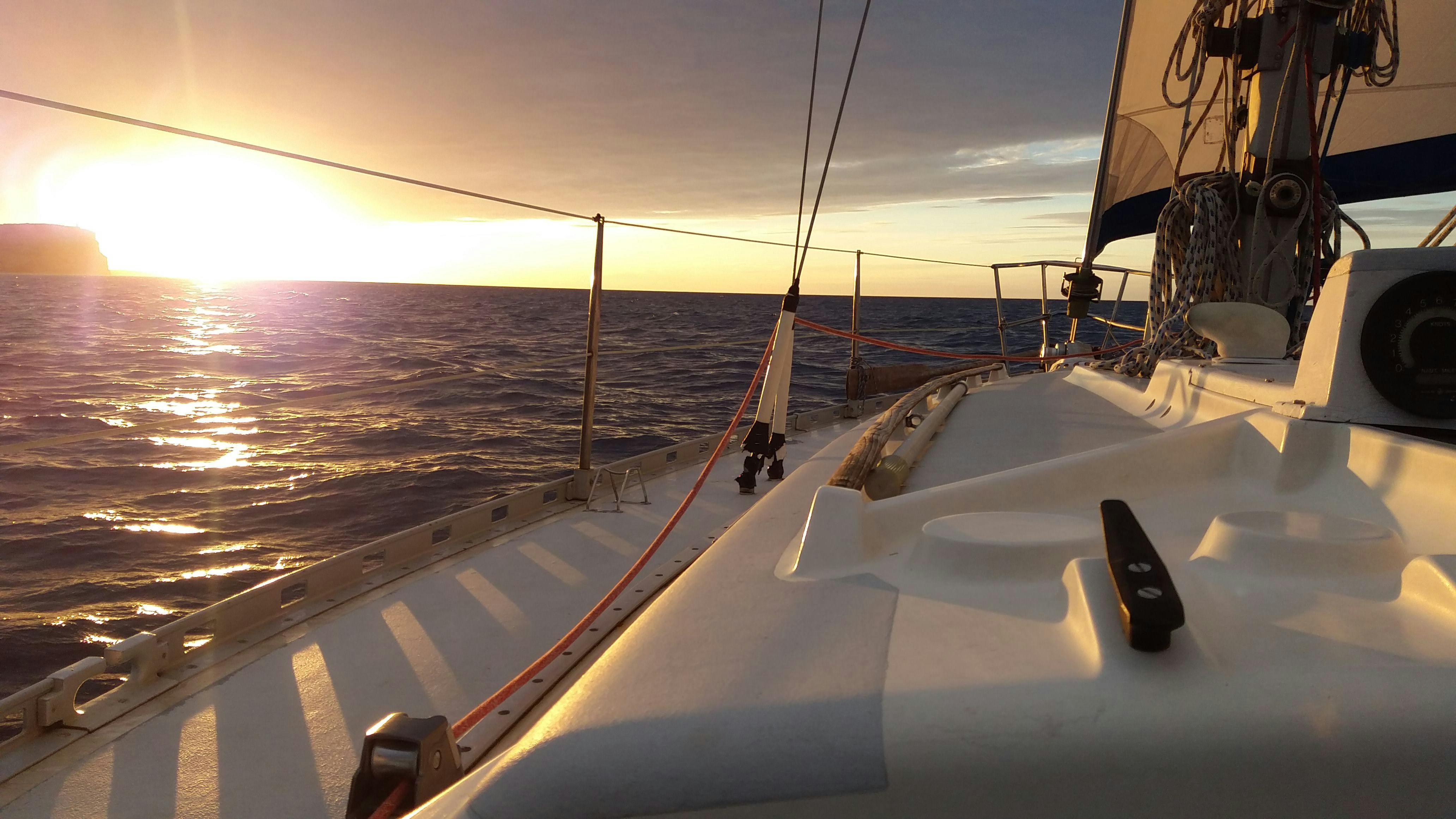 Vue du coucher de soleil depuis le pont d'un voilier sur des eaux calmes, avec le gréement et les voiles visibles.