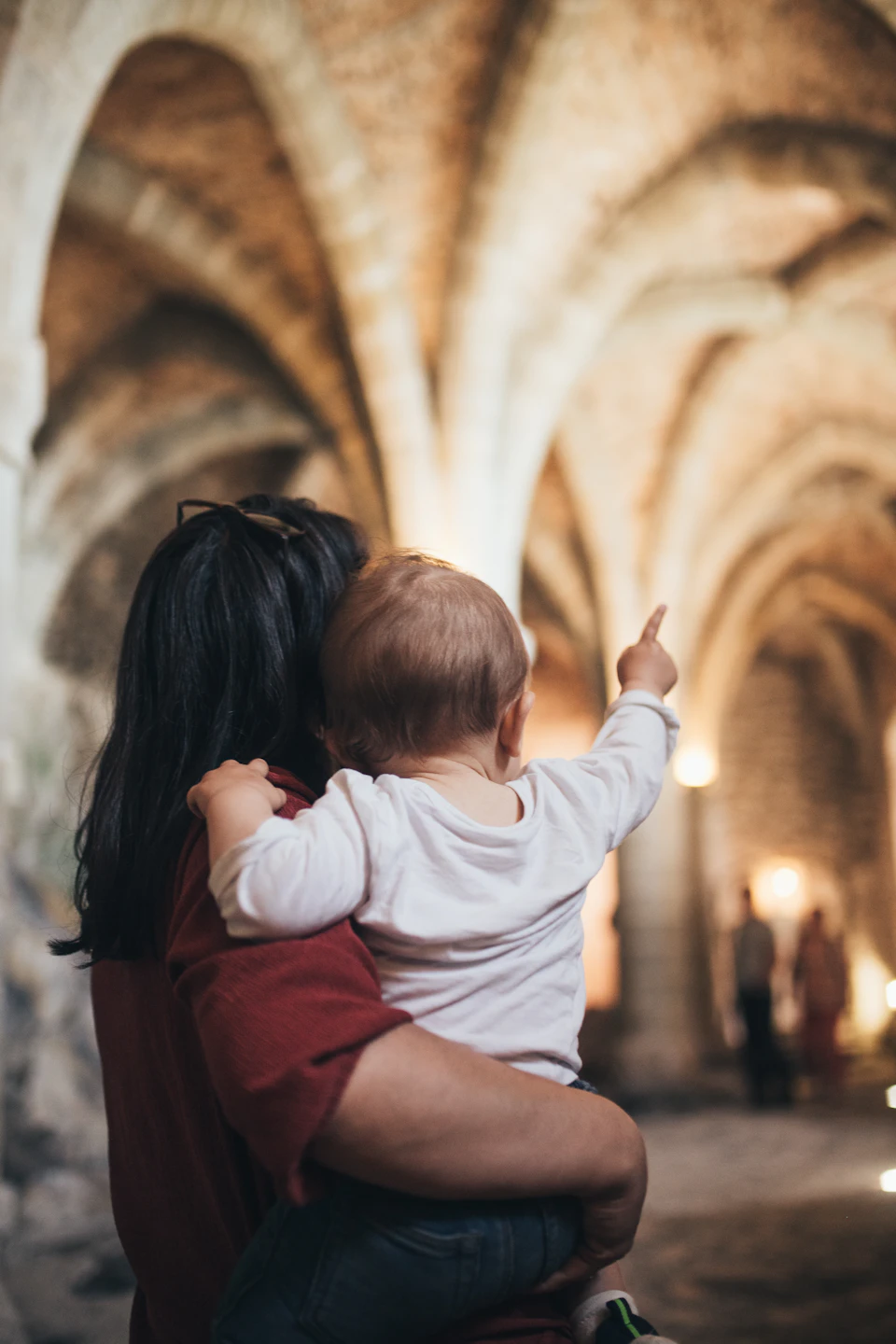 A person holding a baby inside a dimly lit, arched cathedral, with the baby pointing towards something off-frame.