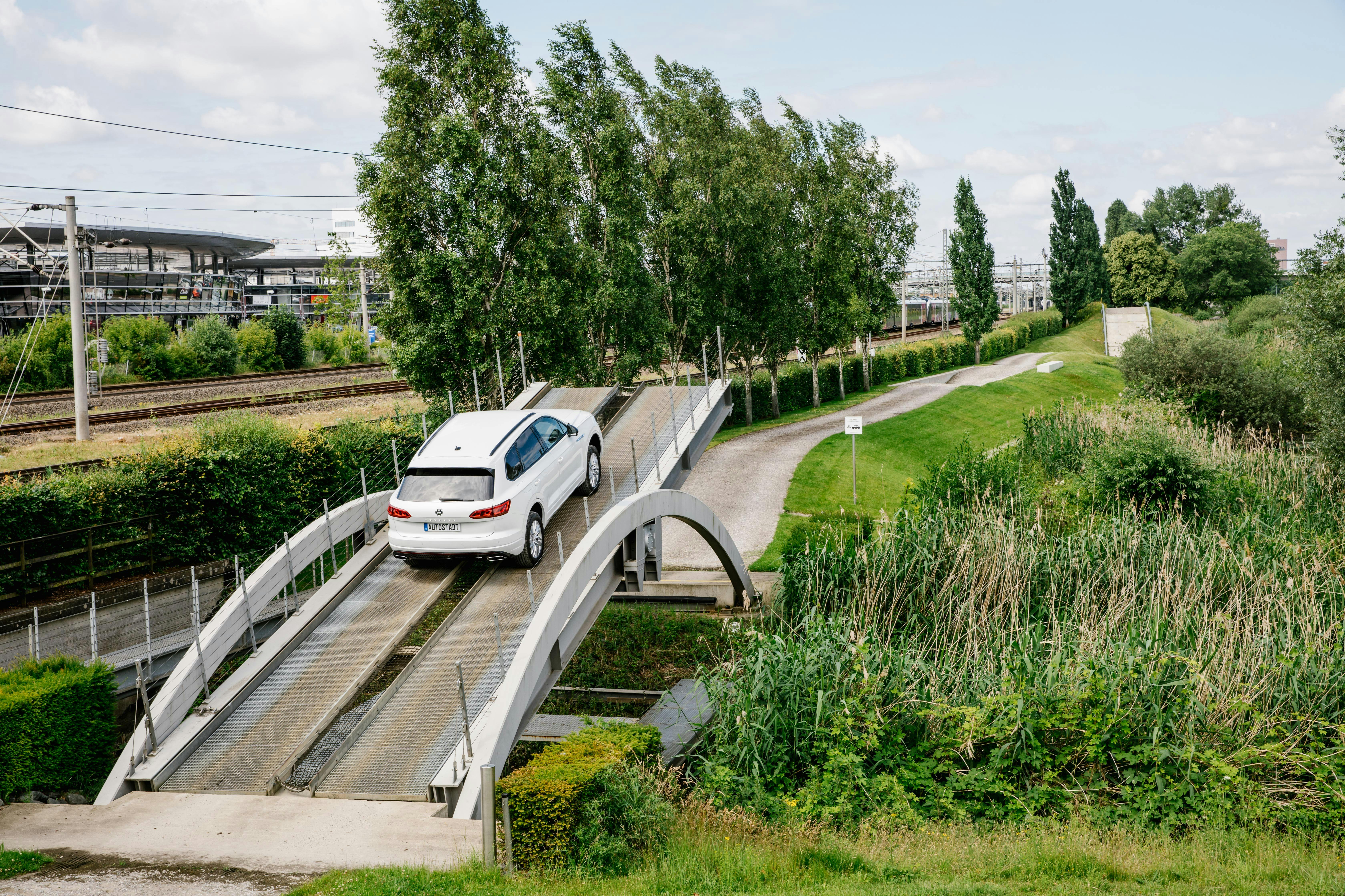 A white SUV drives on an elevated, narrow test track surrounded by greenery and trees near a railway line.