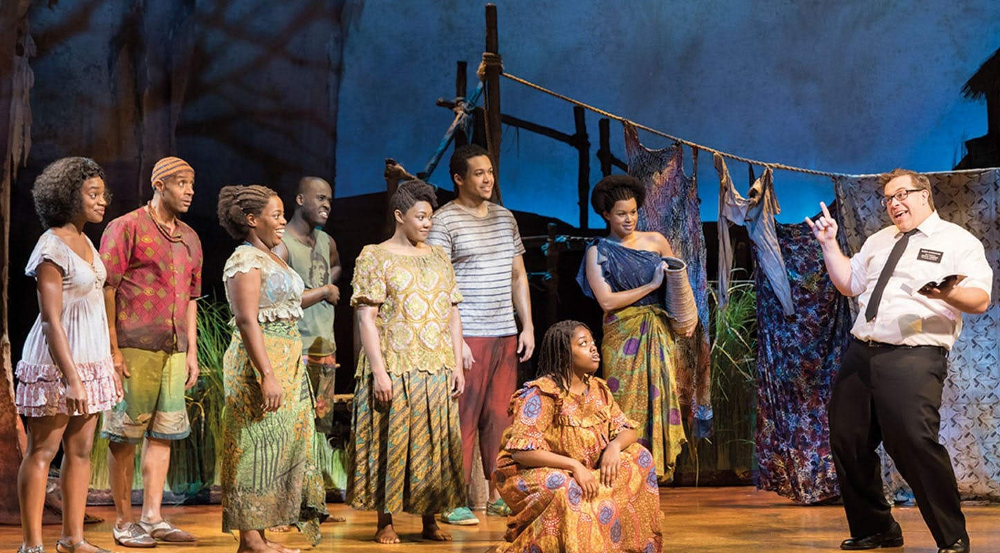 A group of actors in colorful costumes stand and sit on a stage with a blue backdrop, props, and a man in a white shirt gesturing.