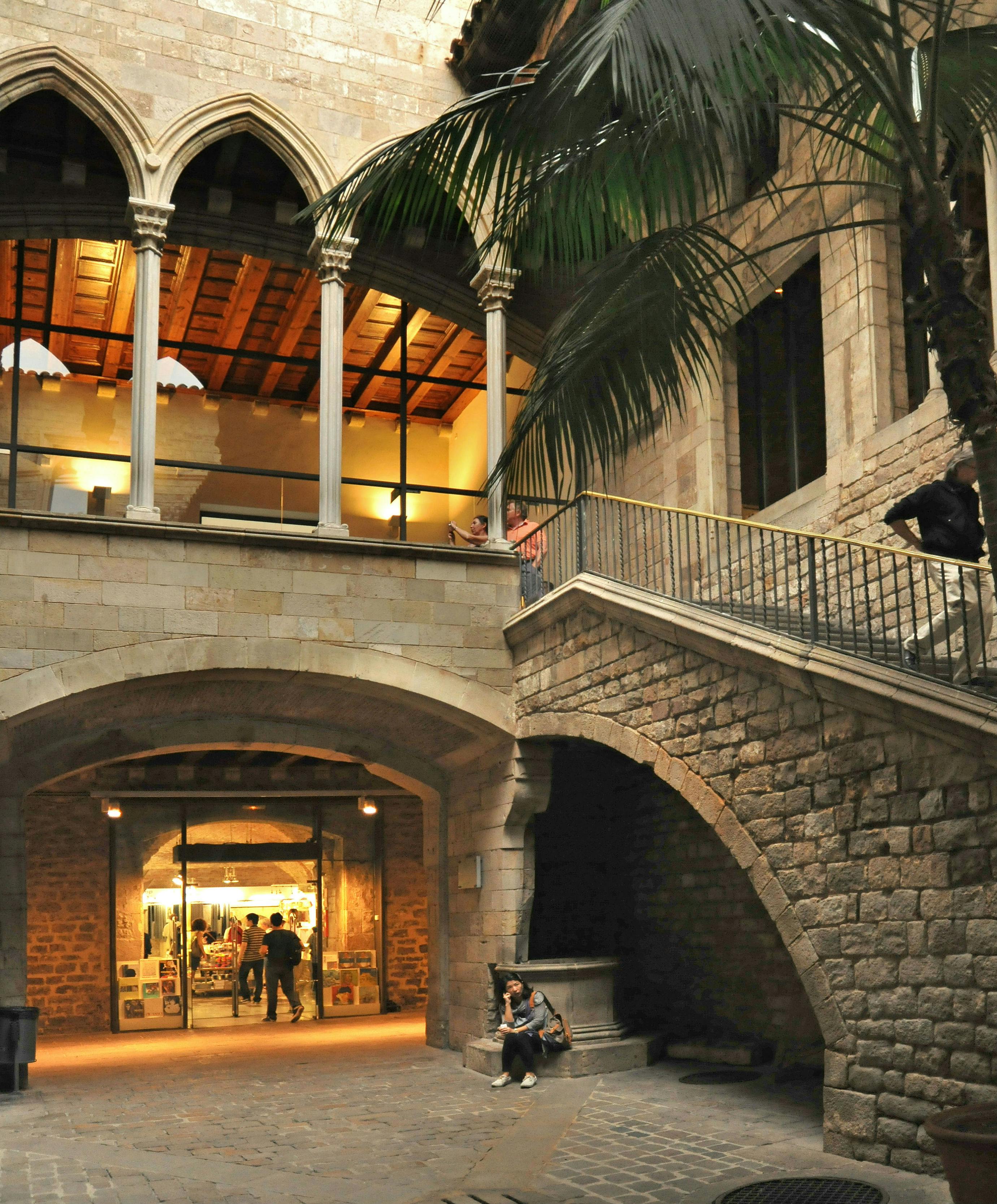 Cour intérieure avec des arches en pierre, un escalier et une galerie au-dessus. Deux personnes sont assises sur un banc en contrebas, d'autres sont visibles à l'intérieur.