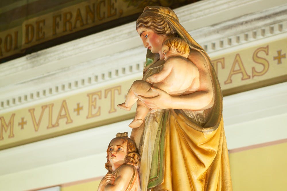Ένα άγαλμα μιας γυναίκας που κρατά ένα μωρό, με ένα άλλο παιδί να στέκεται δίπλα της, με φόντο ένα λατινικό κείμενο.