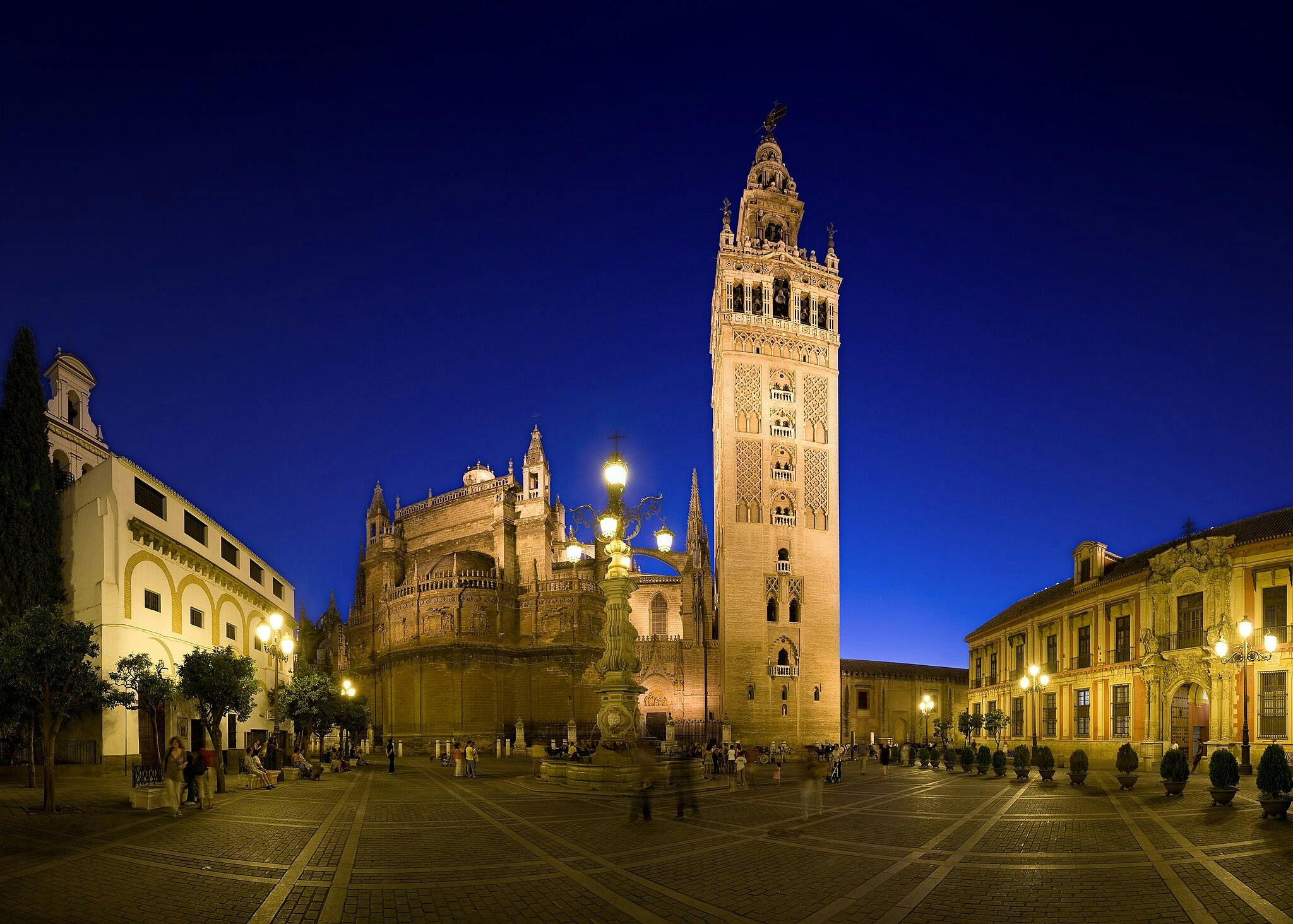 Una grande cattedrale con un alto campanile illuminato di notte, circondata da edifici storici e da una piazza con persone che passeggiano.