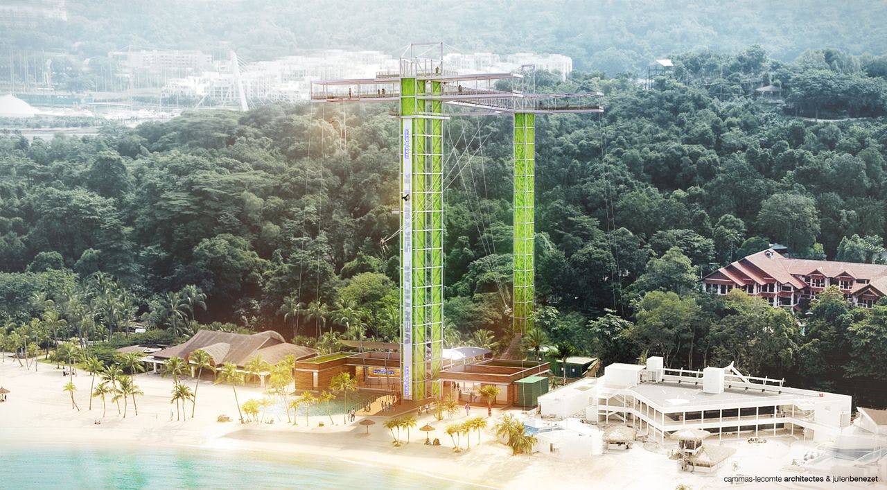 A tall bungee jumping tower with green supports stands on a beach with surrounding tropical forest and nearby buildings.