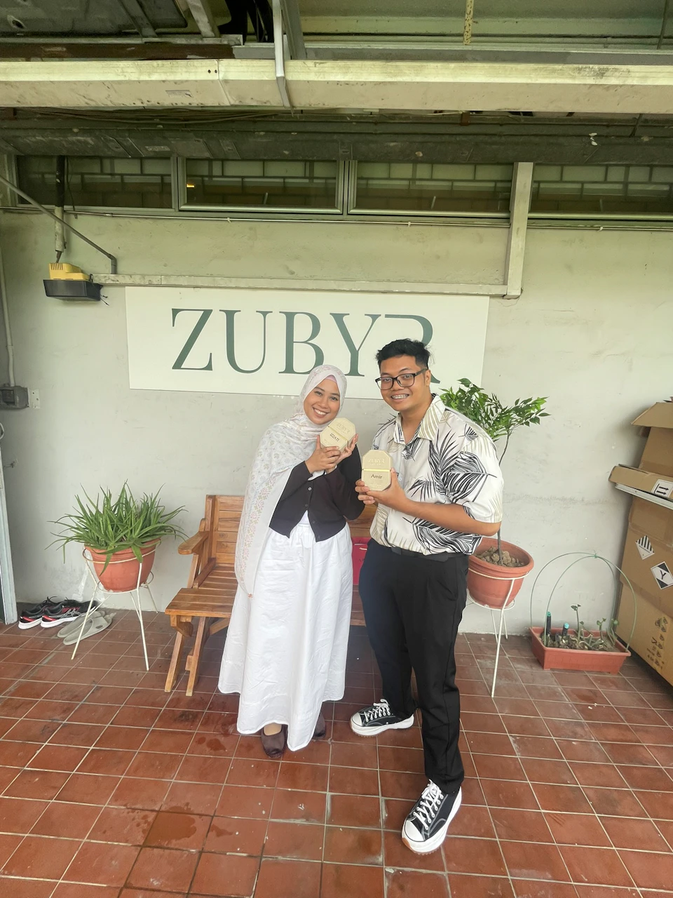 Two people are standing and smiling in front of a sign that reads "ZUBYR." They are holding cups, with plants around them.