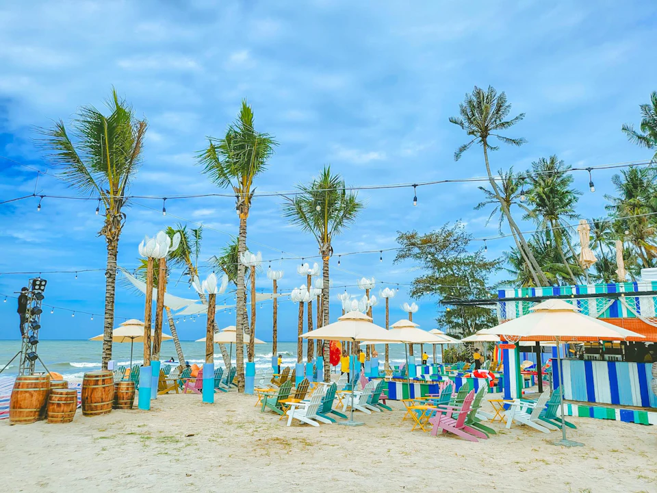A colorful beachside setup with palm trees, chairs, tables, umbrellas, and string lights under a cloudy blue sky.