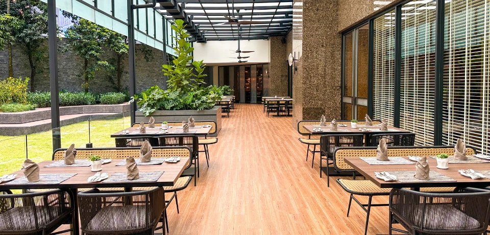 An open-air restaurant with wooden flooring, greenery on the side, and neatly arranged tables and chairs under a covered roof.