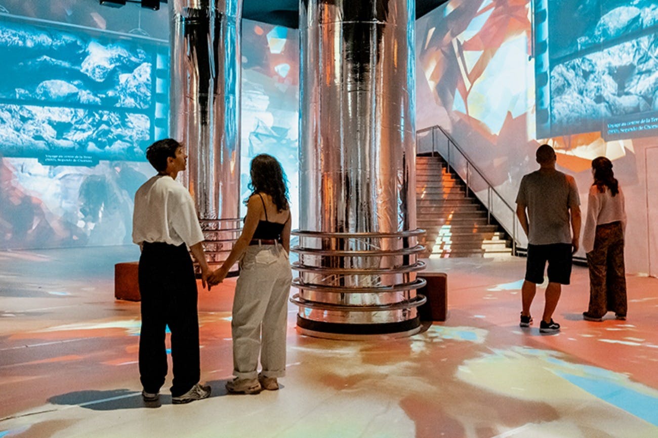 En modern inredning med stora reflekterande pelare, färgglada ljusprojektioner och människor som går eller står.