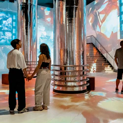 En modern inredning med stora reflekterande pelare, färgglada ljusprojektioner och människor som går eller står.