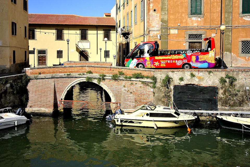 Small boat docked by a brick arched bridge over a canal with colourful tour buses and buildings in the background.