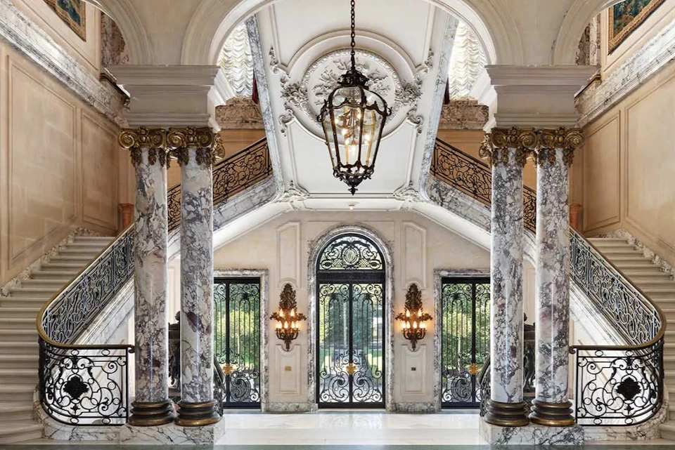 Elegant interior with marble columns, ornate wrought iron doors, chandeliers, and a grand staircase under an arched ceiling.