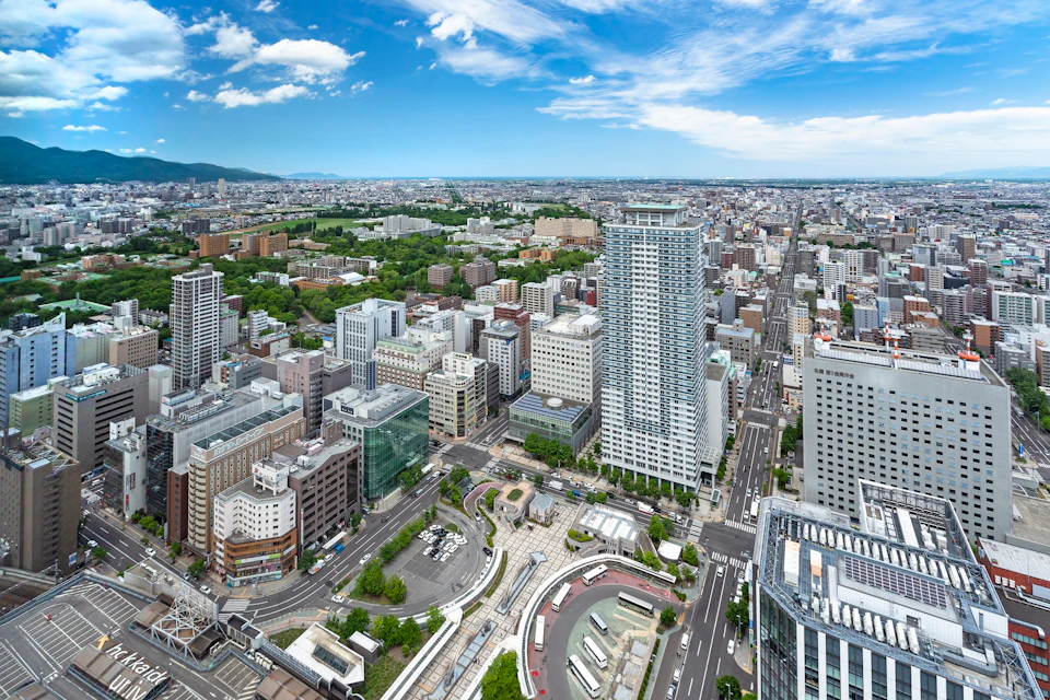 Aerial view of a modern cityscape with tall buildings, roads, green spaces, and a clear blue sky with scattered clouds.