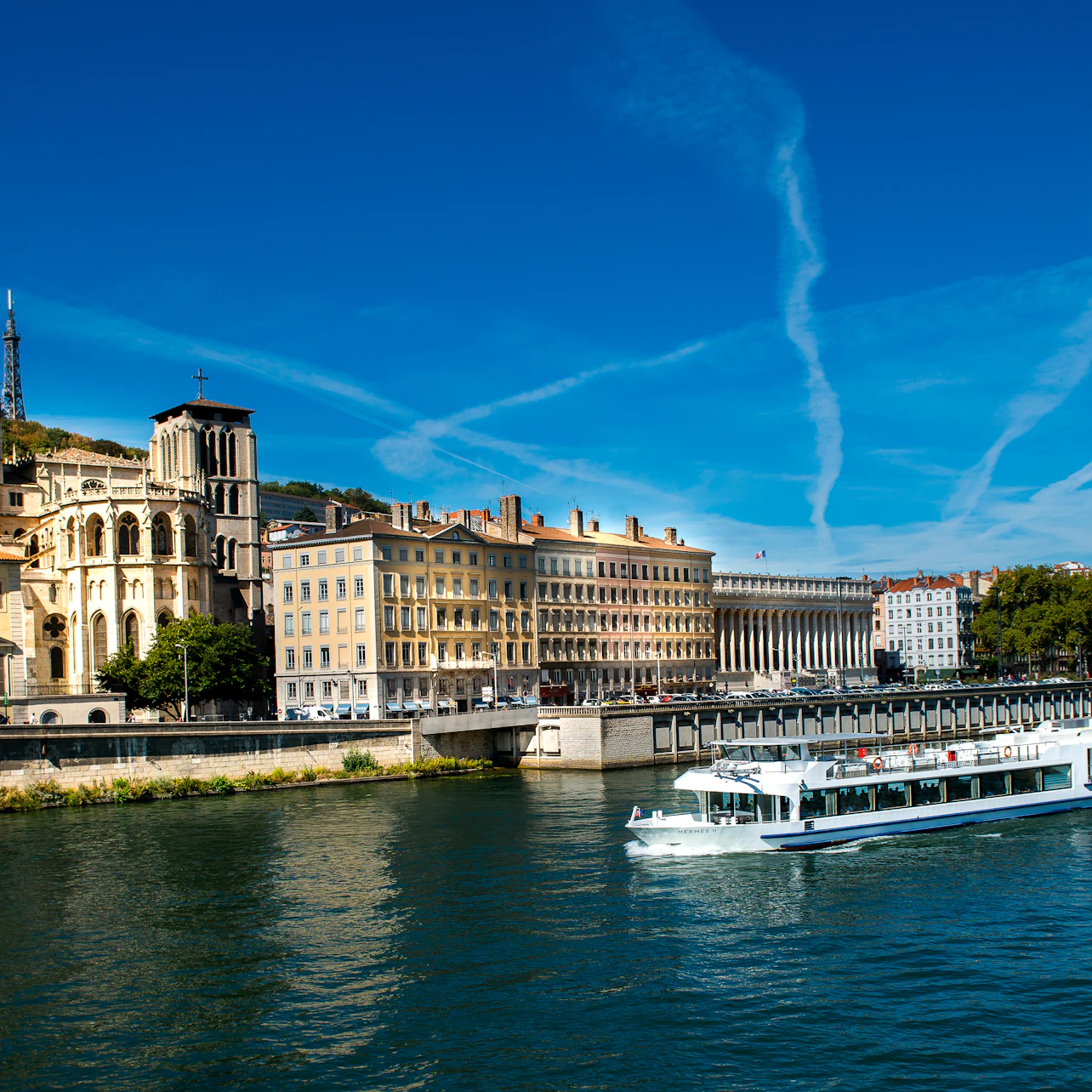 A white boat cruises on a river beside historic buildings under a clear blue sky with white contrails.