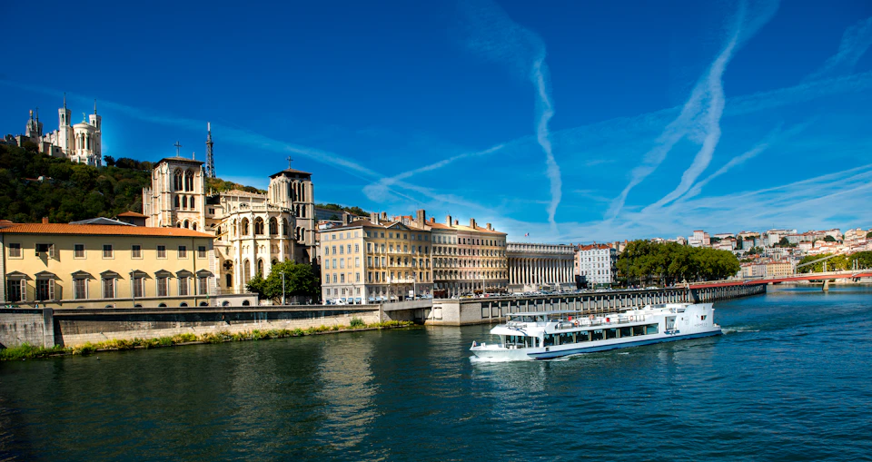 A white boat cruises on a river beside historic buildings under a clear blue sky with white contrails.
