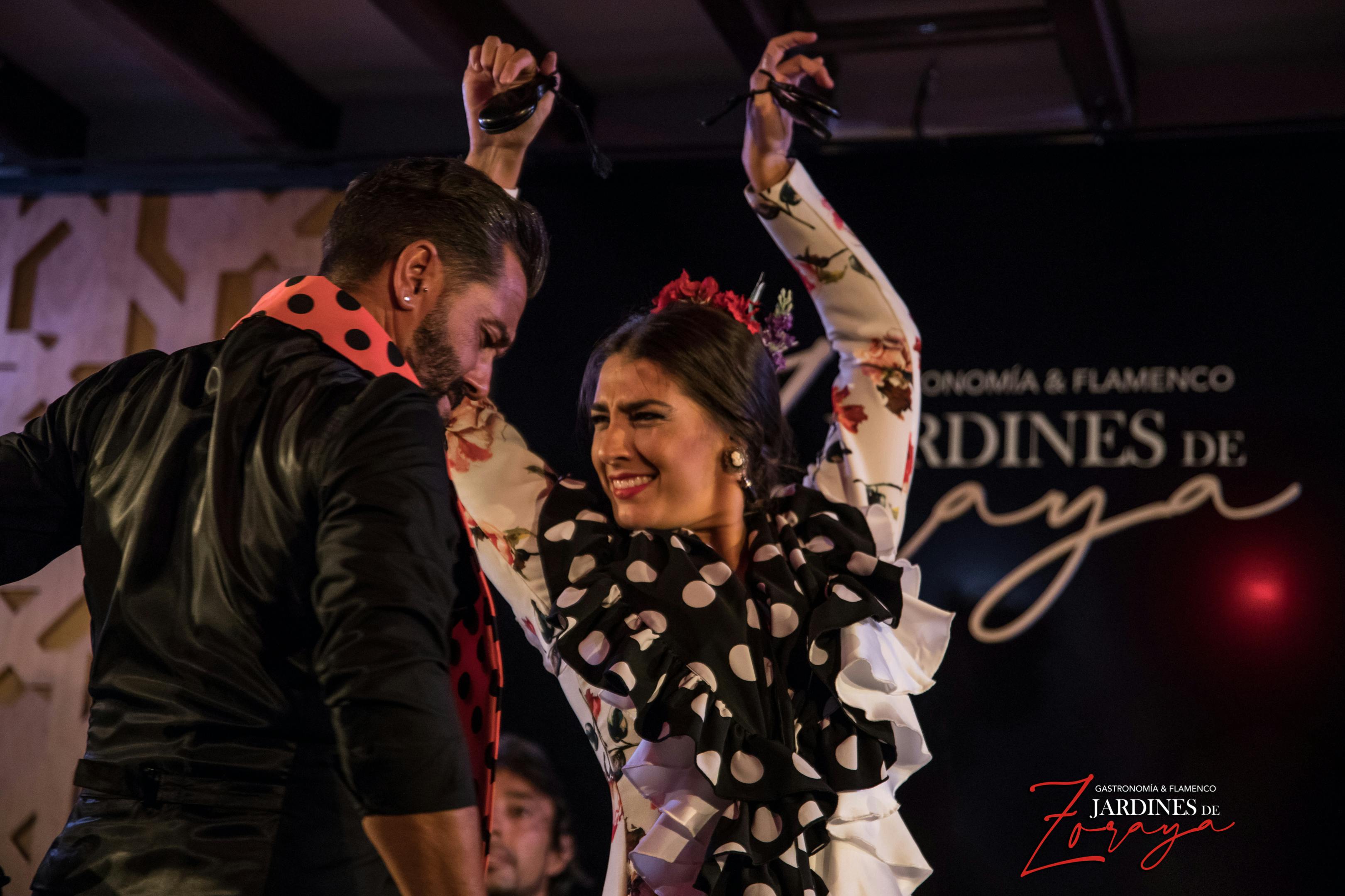 물방울 무늬 드레스를 입고 머리카락을 한 여성이 플라멩코 춤을 추고, 남성이 검은 셔츠와 물방울 무늬 스카프를 두르고 있습니다.