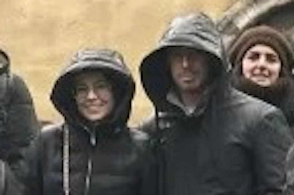 A group of people in warm clothing standing in front of a stone building with tall arched windows on a cloudy day.