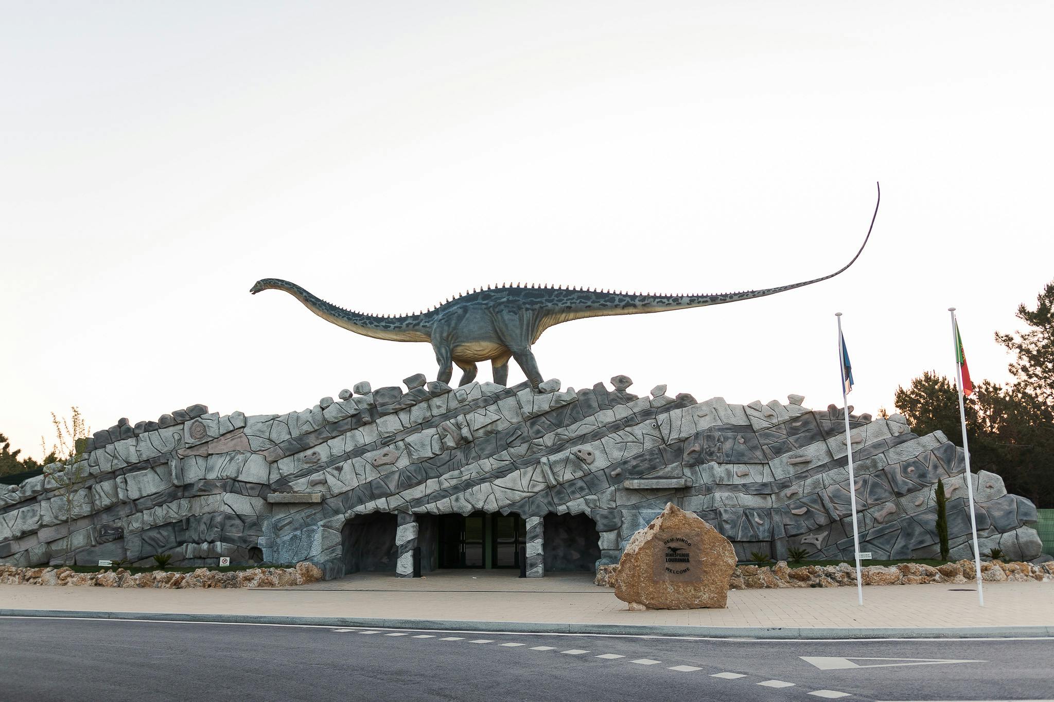 Grande sculpture de dinosaure au sommet d'un bâtiment en pierre avec des entrées arquées, un mât de drapeau et un rocher à l'avant.