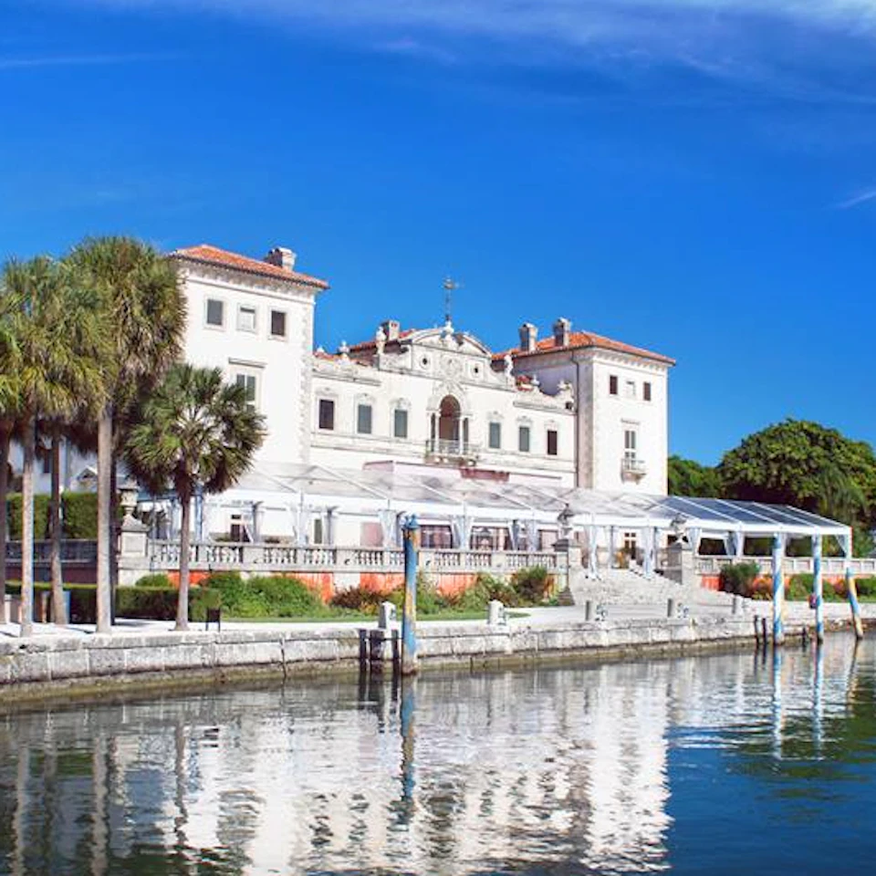 A grand, historic building with white walls and orange roofs, surrounded by palm trees and adjacent to a calm reflective waterway.