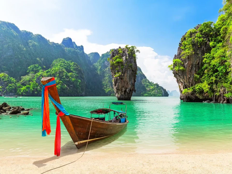 A long-tail boat with colorful ribbons is anchored on a sandy shore, with tall, lush cliffs and green water in the background.
