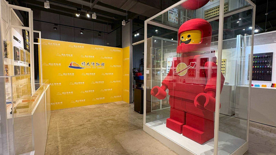 A large red LEGO-style astronaut figure displayed in a glass case, with a yellow wall featuring a logo in the background.