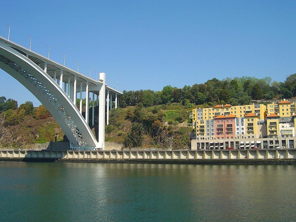 En stor bro korsar över en flod med färgglada byggnader och en skogsklädd kulle i bakgrunden under en klarblå himmel.