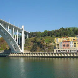 En stor bro korsar över en flod med färgglada byggnader och en skogsklädd kulle i bakgrunden under en klarblå himmel.