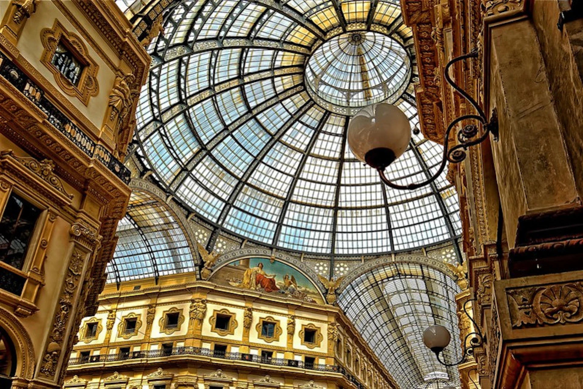Ornate glass-domed ceiling with intricate metal framework inside a grand, historic building. Chandeliers and decorative details are visible.
